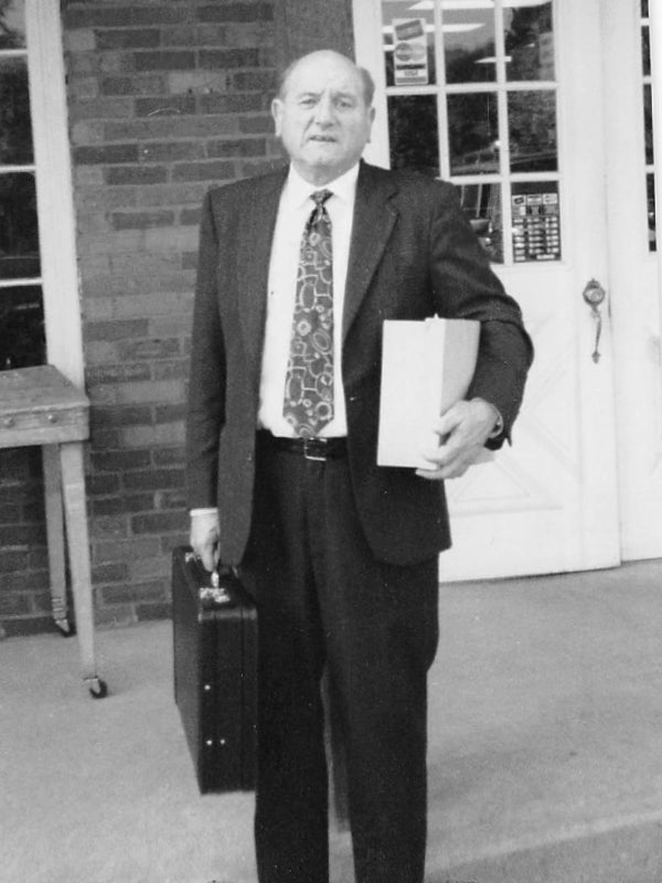 1992. Hank in a suit holding a briefcase and a box outdoors. Text below the image: Hank passed away in 1992, but the legacy he built continues to shape who we are today. He left a mark on everyone who met him—known for his tireless work ethic, firm handshake, and deep love for his family. Hank didn’t just build a business; he built a life rooted in integrity, grit, and the values that still guide us.