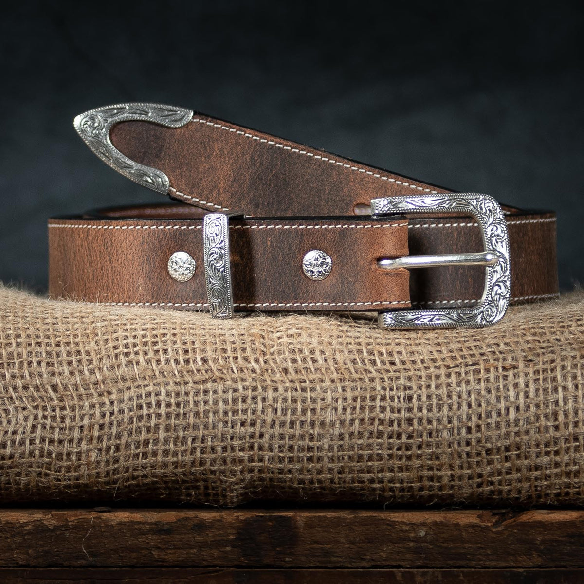 The Willow Bison by Hanks Belts, a women's brown leather western belt with a floral buckle and ornate silver accents, is coiled on burlap against a dark backdrop and wooden surface.