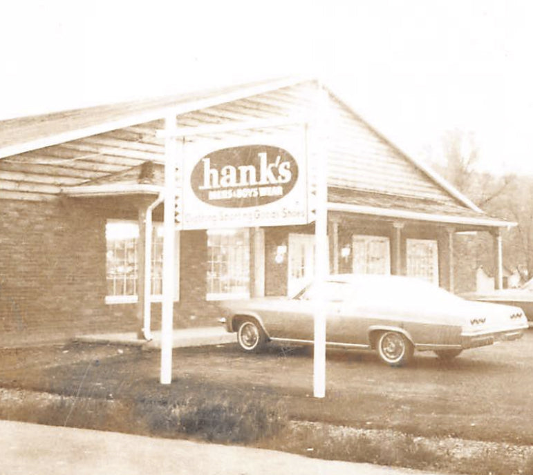 Mid 60's .Vintage photo of a building with 'hank's' sign and a car parked in front. Text below the image: With the growth of the business and working 12-14 hour days, sights were set on expansion into a new facility. A new building and addition a year later gave room to expand his offerings.