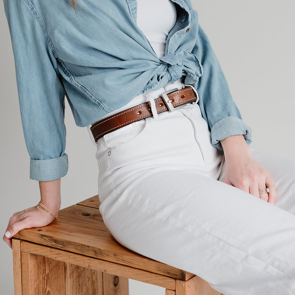 A person sits on a wooden stool, wearing a light denim shirt tied at the waist, a white top, high-waisted white jeans, and The Bonnie - 1.25" belt from Hanks Belts. Their left arm rests on the stool, showing a bracelet and rings.
