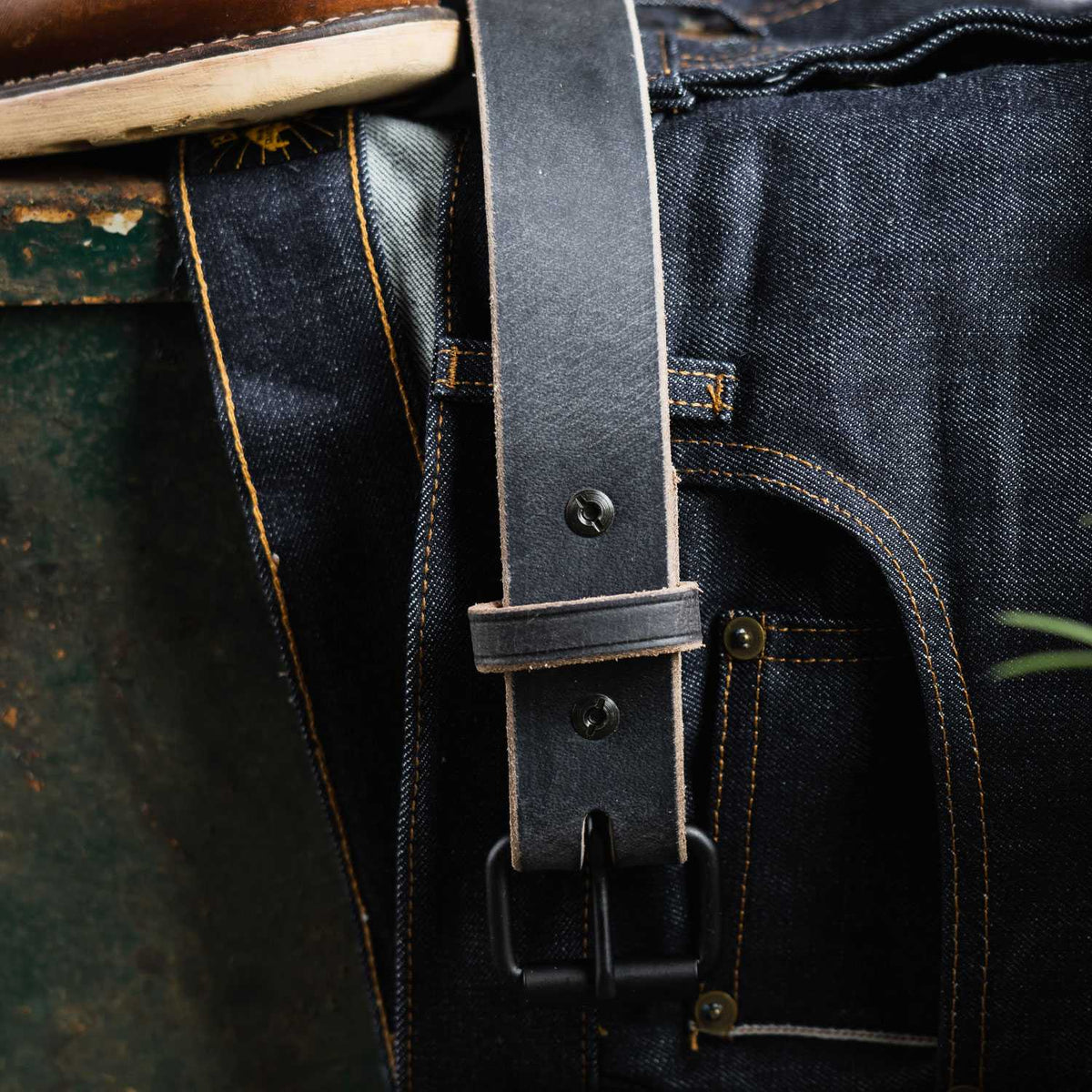 A close-up of The Glacier Grey Retro Style by Hanks Belts, crafted from Water Buffalo leather, looped through dark blue denim jeans with contrast stitching beside a distressed green metal surface.