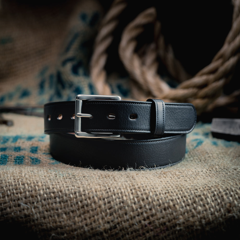 The Tuscan Embossed Lined by Hanks Belts, a premium English Bridle Leather belt with a stainless steel buckle, is coiled on burlap fabric with rustic décor and thick rope softly blurred in the background.