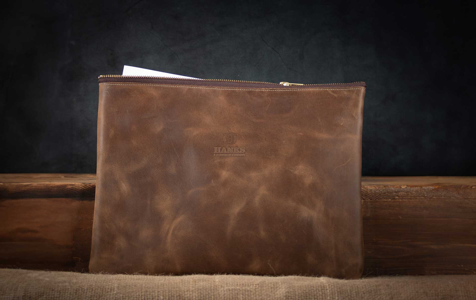 A brown leather zippered pouch with a subtle embossed logo sits upright on a wooden surface, with a white sheet of paper partially visible inside, against a dark background.