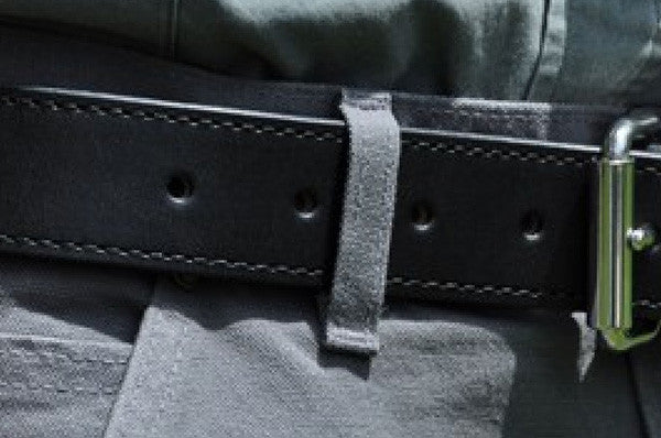 Close-up of a black leather belt with visible stitching, fastened around gray pants. The belt has several holes and a silver buckle is partially visible on the right side.