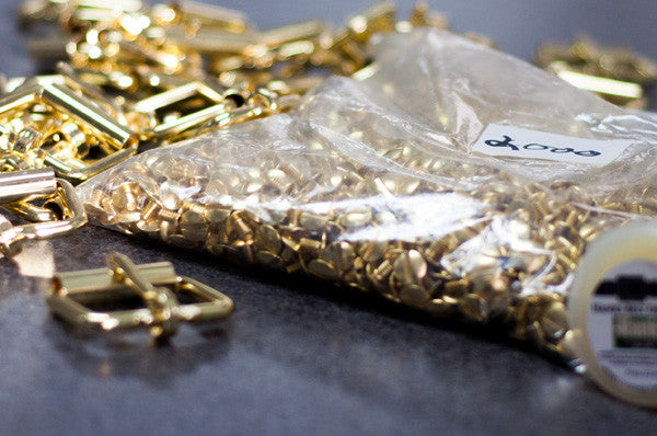 A clear plastic bag labeled "2000" filled with small gold-colored metal clasps sits on a table, with larger gold hardware pieces and another container nearby.