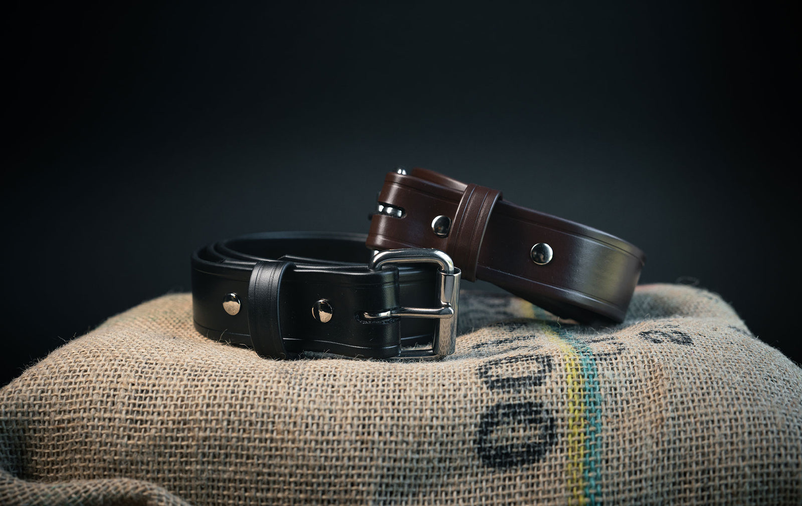 Two leather belts, one black and one brown, are rolled up and placed on a burlap sack. The belts have metal buckles and hardware, and the background is dark, emphasizing the belts.