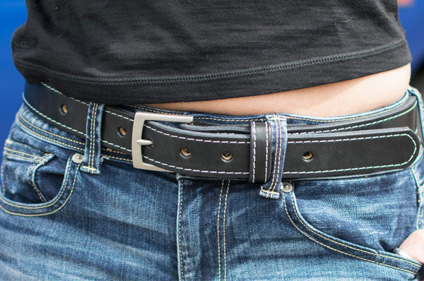 A person wearing blue jeans and a black belt with white stitching, paired with a black shirt. The belt is fastened and goes through the belt loops of the jeans.