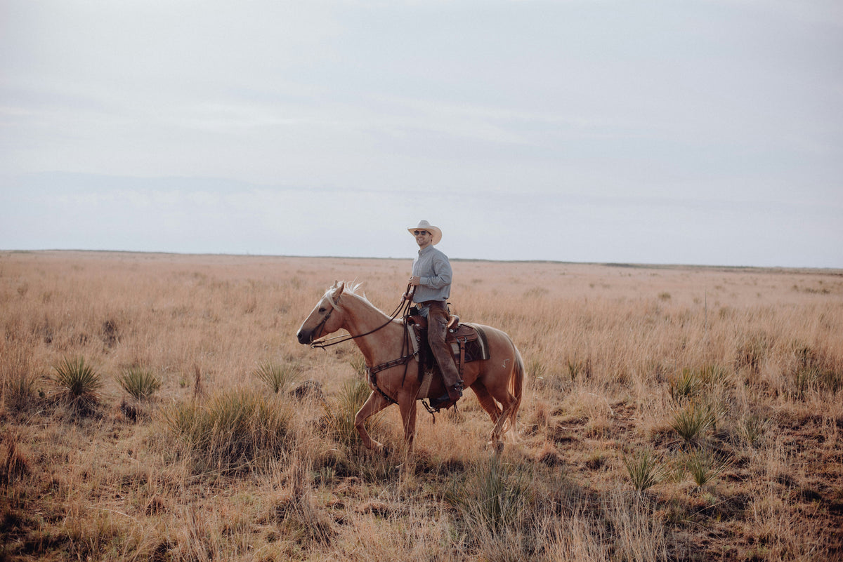 A cowboy wearing a hat rides a horse through a dry grassy plain under a clear sky.