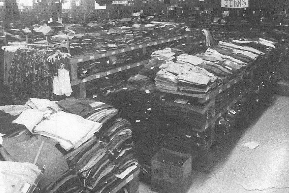 Mid 50's .Black-and-white photograph of a clothing store interior with long tables stacked high with neatly folded garments, including pants and shirts, arranged in rows; clothing racks and signage are visible in the background. Text below the image: Hank purchases the business and continues with military surplus but starts to add more clothing and sporting goods, making the business his own. 