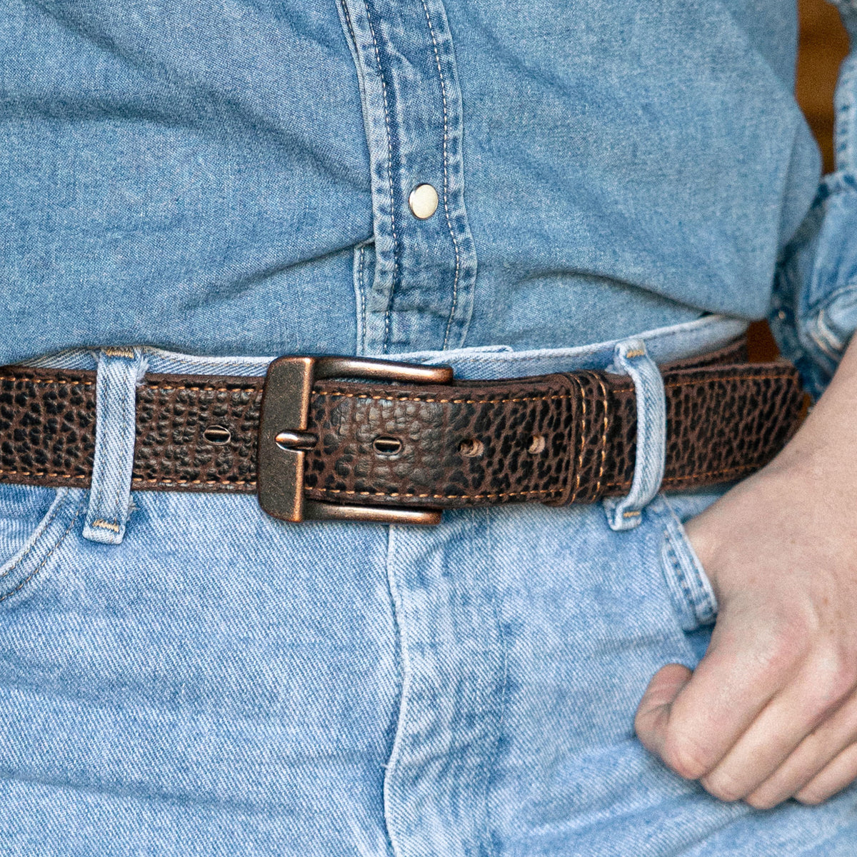 A close-up of a person in a blue denim shirt tucked into light blue jeans, wearing The Yellowstone belt by Hanks Belts at the waist, with their right hand resting on their hip.