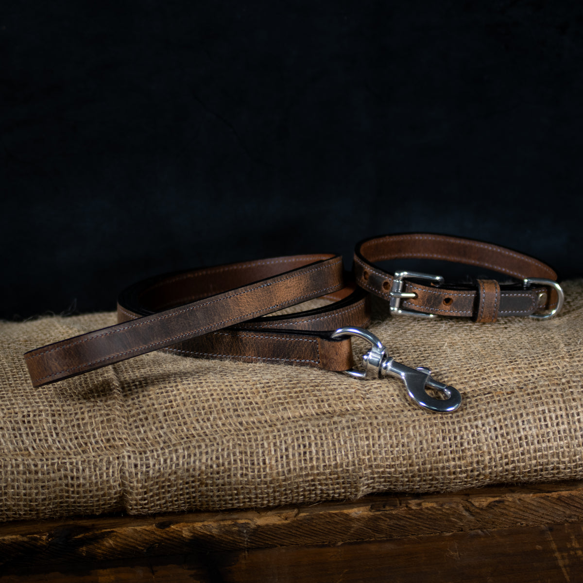 The Hanks Belts Ollie Bison Collar in brown, paired with a matching leash and stainless steel hardware, is displayed on burlap against a dark background.