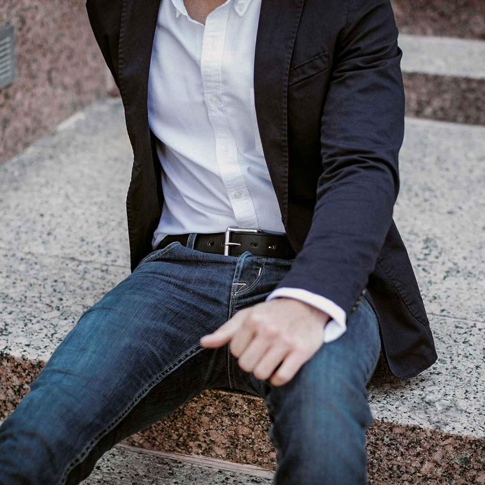 A person in a black blazer, white shirt, blue jeans, and The Tuscan Embossed Lined belt by Hanks Belts with a stainless steel buckle sits on stone steps. The photo is cropped above the mouth, showing only the lower face and torso.